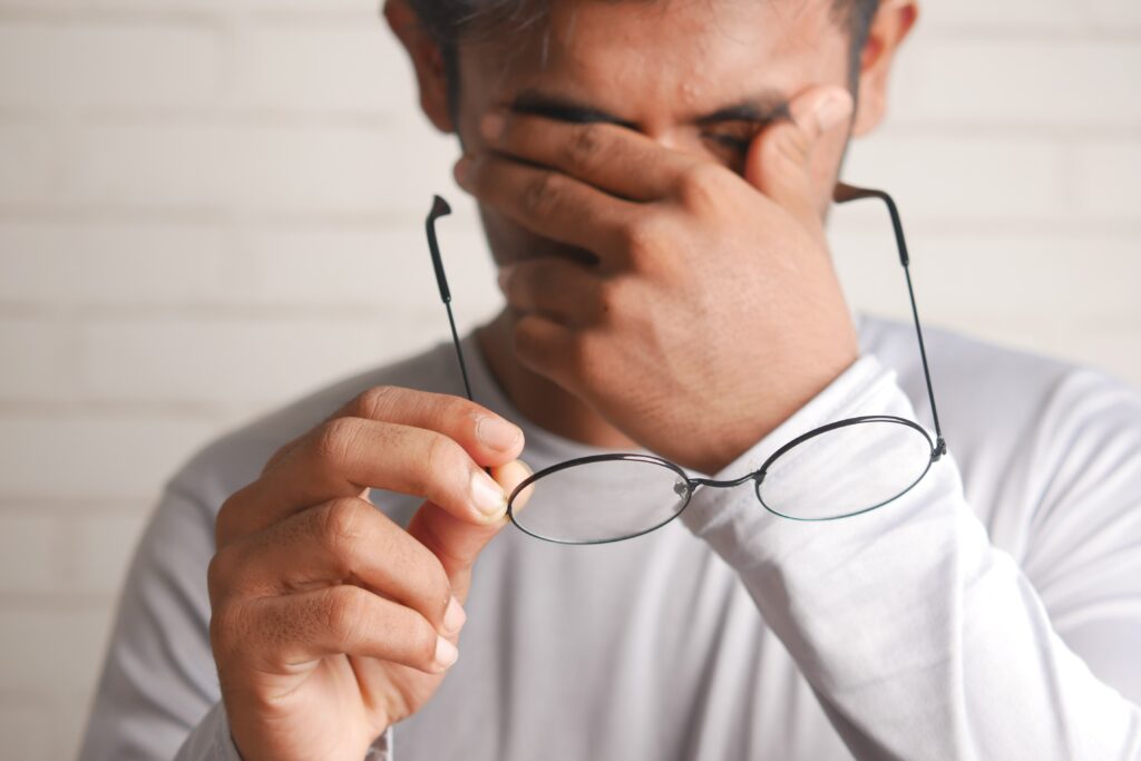 una persona masculina con unas gafas de ver en la mano derecha y con los ojos tapados con la mano izquierda